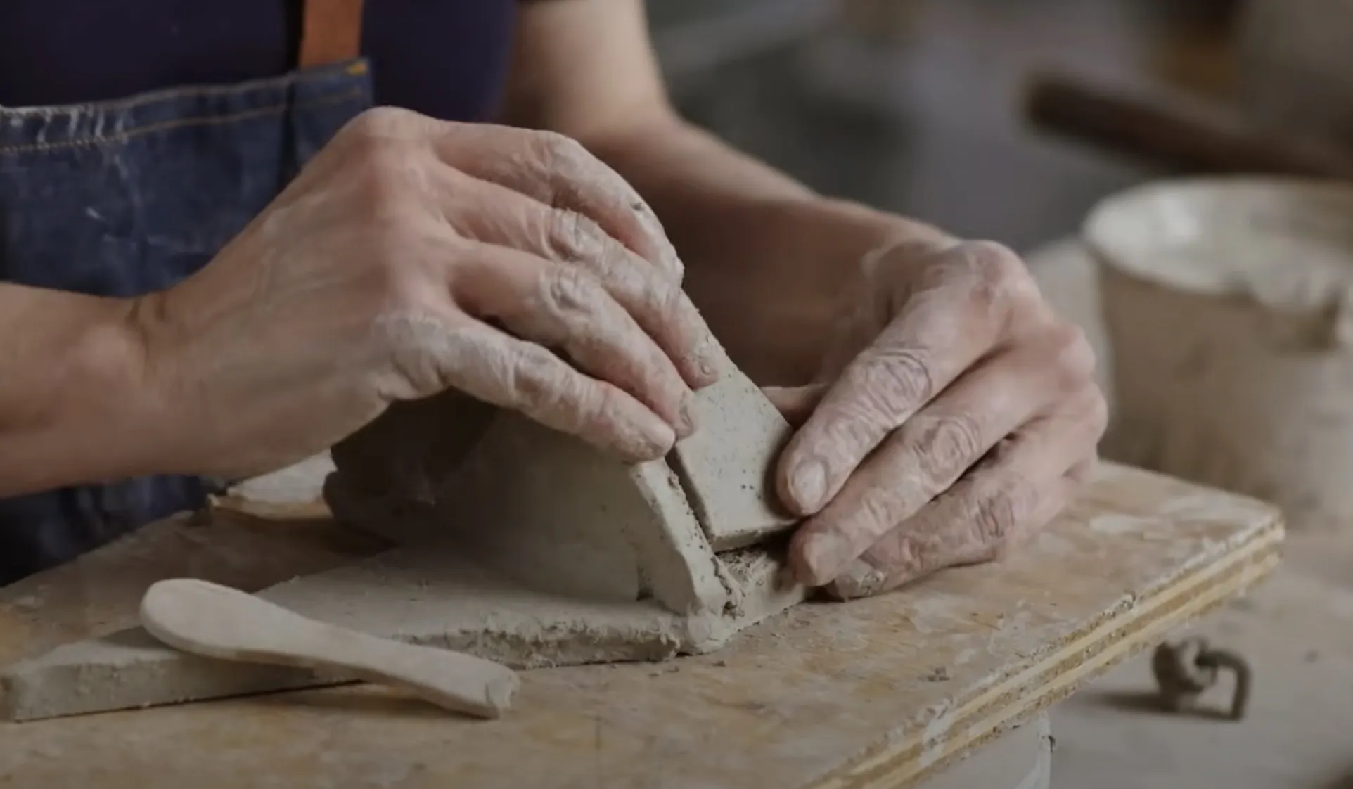 A potter's hands pressing and shaping a rectangular block of grey clay on a wooden workboard, with a wooden modelling tool resting nearby.